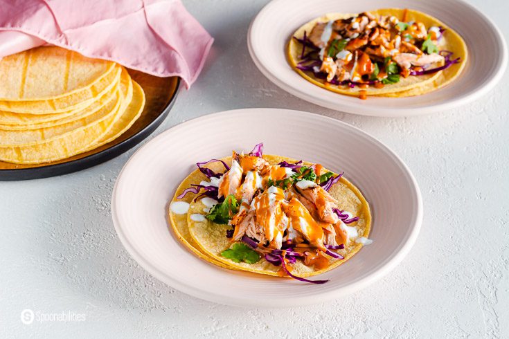 Two plates with fish tacos and a tray with corn tortillas on the left top side, The fish selected for those tacos is salmon, which was cooked in the air fryer. Recipe at Spoonabilities.com