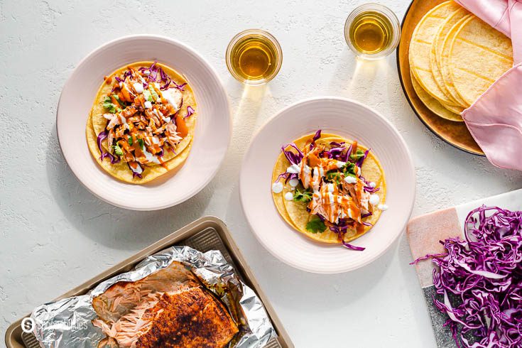 Overhead photo with two pink plates with air fryer salmon tacos. On the top right side is a plate with corn tortillas, next to that plate shredded red cabbage, and on the left bottom side a tray with the salmon cooked in the air fryer. Recipe at Spoonabilities.com