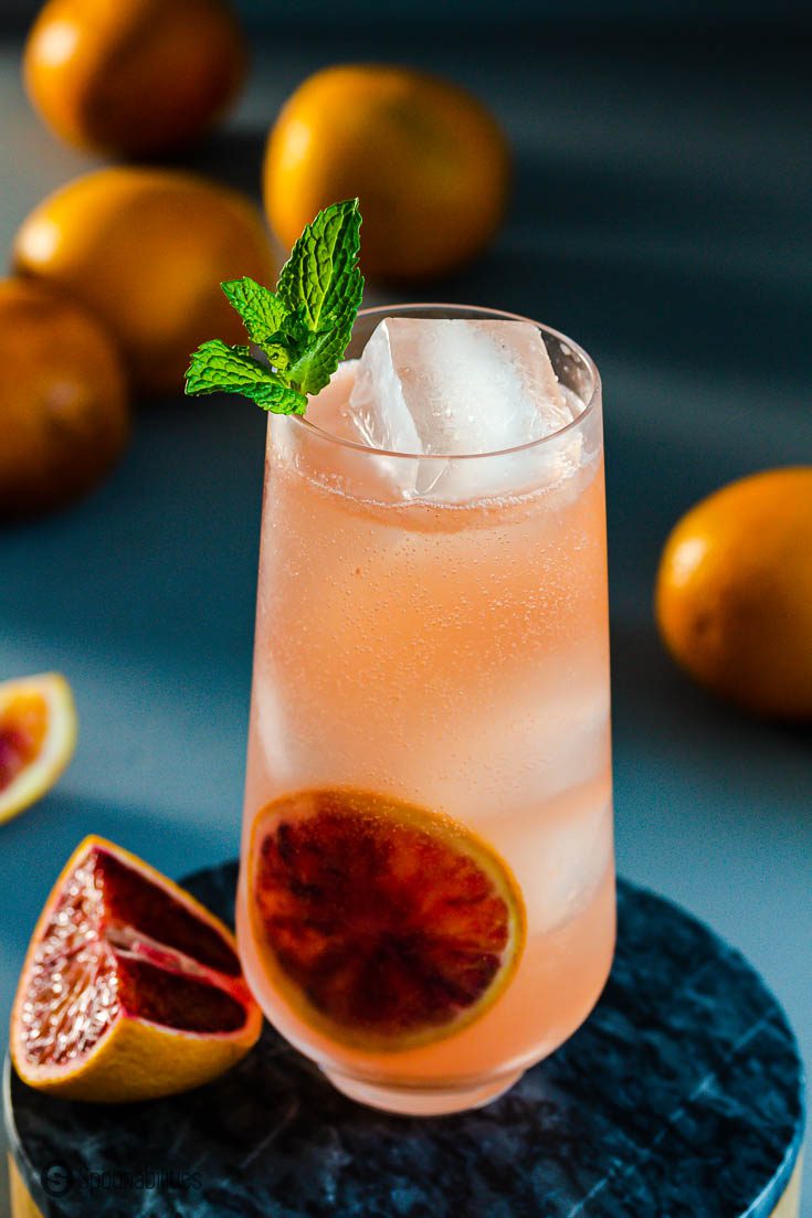 A tight close-up photo of a blood orange cocktail in a tall glass and near the glass a wedge of fresh blood orange. Recipe of this cocktail available at Spoonabilities.com