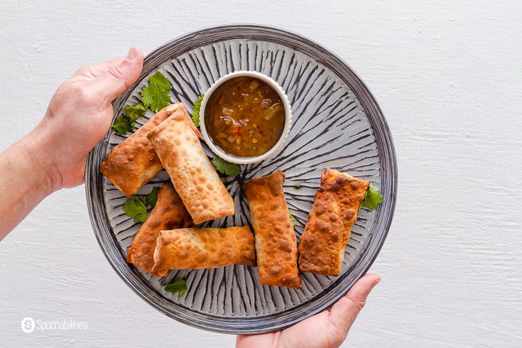 Two hands holding a grey plate with some egg rolls stuffed with coleslaw veggie mix cooked in the air fryer until crispy perfection. These egg rolls are served with roasted pineapple habanero sauce. Recipe at Spoonabilities.com