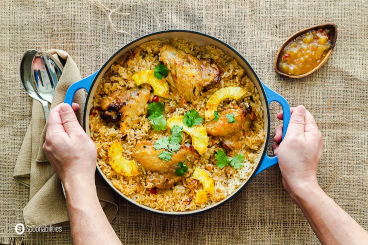 Two hands hold the cast-iron casserole with rice and chicken seasoned with a whole bottle of roasted pineapple habanero sauce by Terrapin. Recipe at Spoonabilities.com