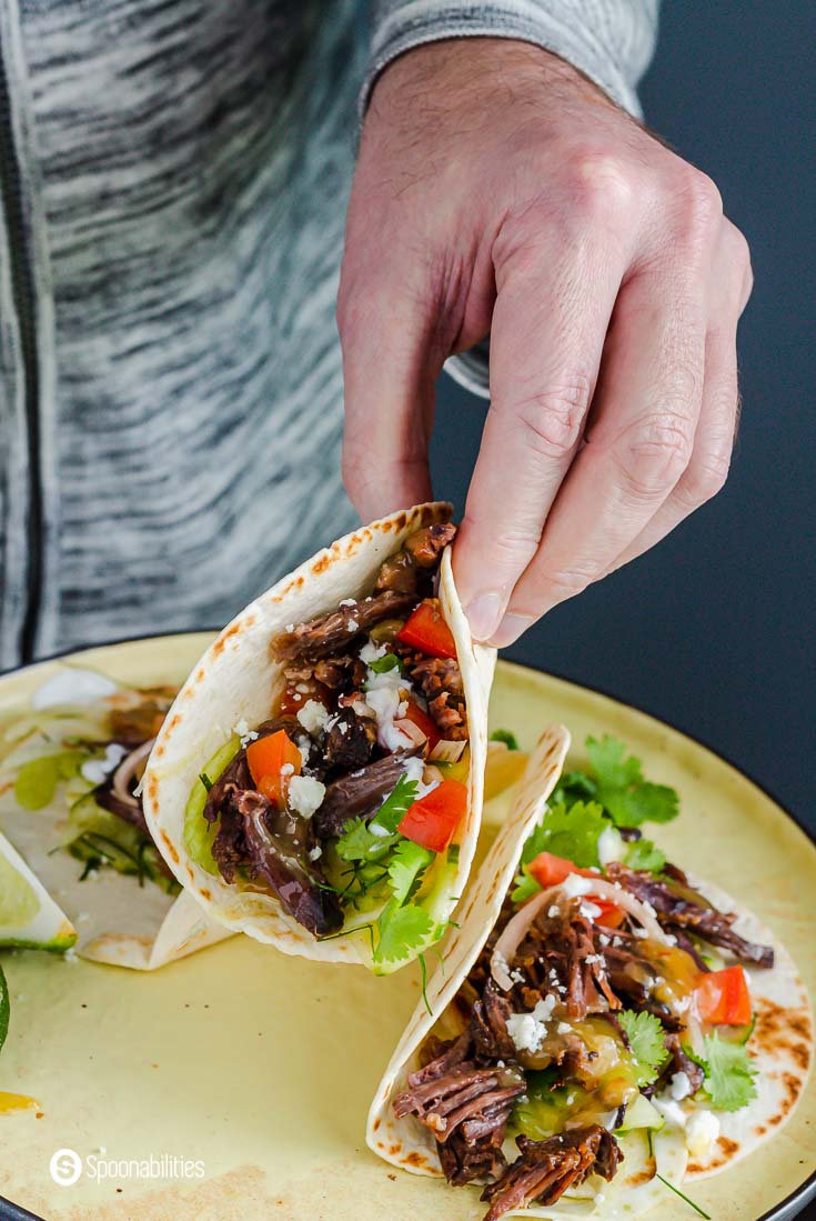 A hand holding one instant pot short ribs tacos with tropical tequila sauce. The other hand is holding the plate with two more tacos. Recipe at Spoonabilities.com