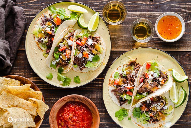 Overhead photo with two plates with three short ribs tacos, each a bowl with chips and salsa. Also, two glasses of beers and a small bowl on the right top corner with the Tropical Tequila Sauce by Terrapin Ridge Farms. Recipe at Spoonabilities.com