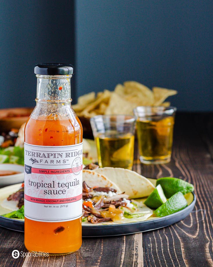 A bottle of Tropical Tequila Sauce and in the background a plate with tacos and some beers. recipe at Spoonabilities.com