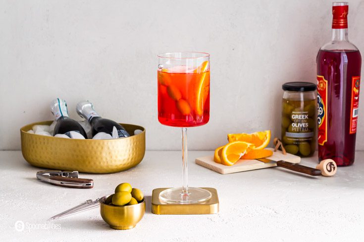 The Italian summer cocktail served in a glass wine. In the background a bottle of prosecco, Green Greek olives, and the aperitif Aperol. Recipe at Spoonabilities.com