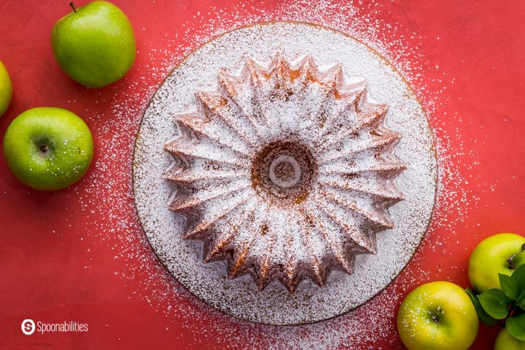 Overhead photo with an olive oil cake on a round plate, The cake has powdered sugar and some Granny Smith apples around the picture frame. Recipe at Spoonabilities.com