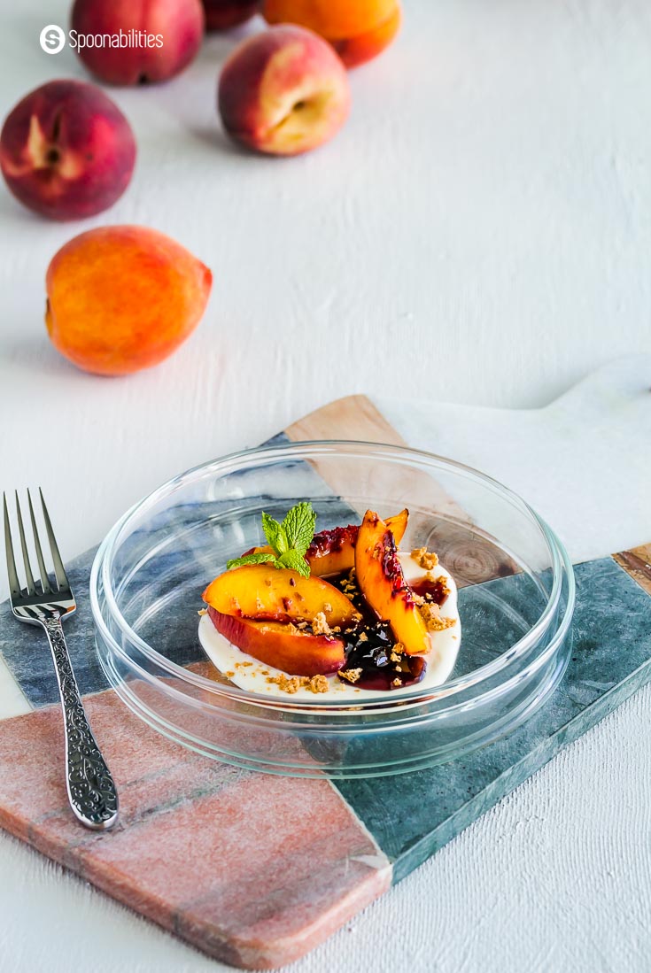 A glass plate on top of a marble board with a peach dessert on the plate with baked peaches, black cherry preserves, and Creme Fraiche. Recipe at spoonabilities.com