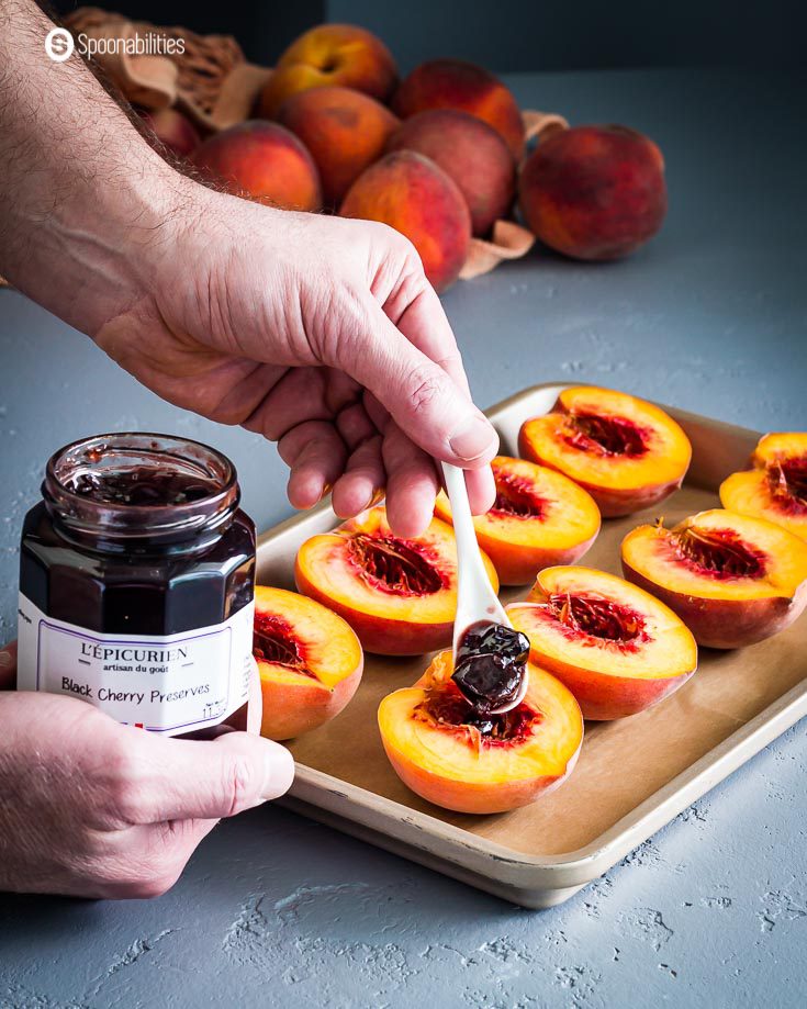 A baking tray with peaches cut in halves and a hand with a jar of black cherry preserves, and the other hand with a spoon putting the jam on the peach. Recipe at spoonabilities.com