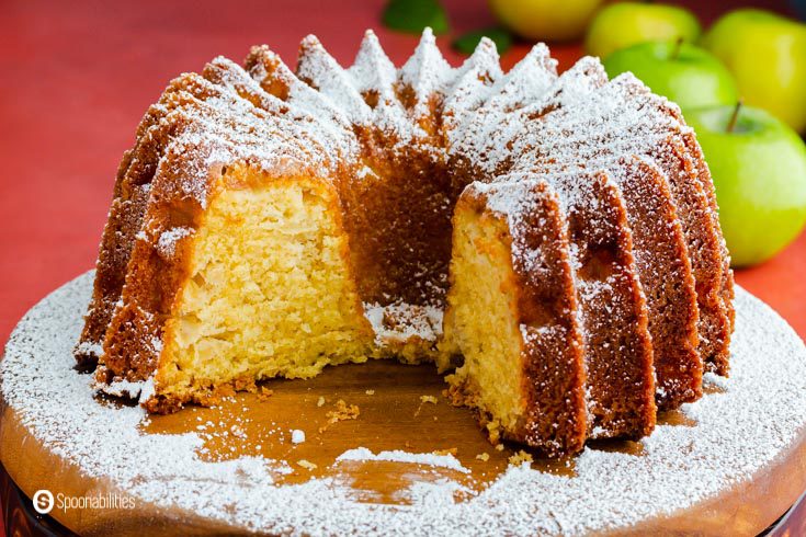 A bundt cake with a piece out, and it's showing the inside crumb of the cake. The cake is made out of olive oil and apples. Recipe at Spoonabilities.com