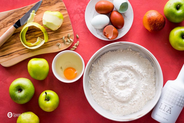 A tabletop with the apple and olive oil cake ingredients, Some of the ingredients are LIA Extra Virgin Olive Oil, Granny Smith Apples, eggs, orange, cardamon, and a flour mixture in a bowl. Recipe at spoonabilities.com