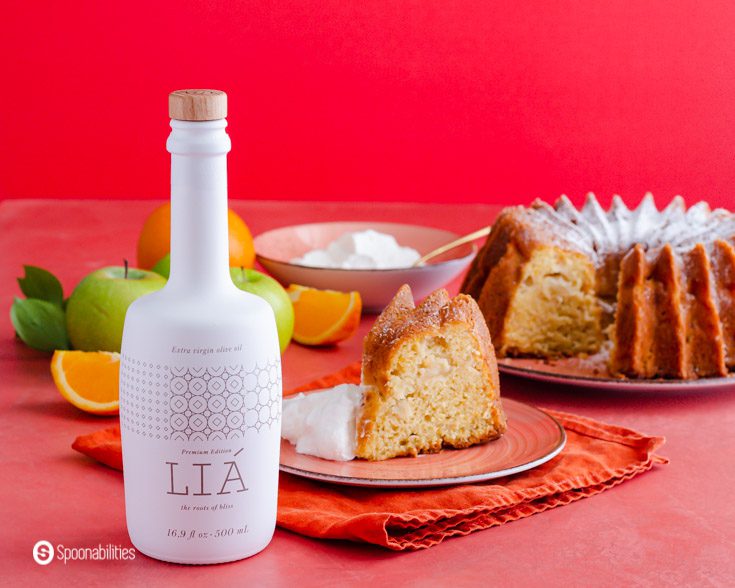 A beautiful Lia Extra Virgin Olive oil bottle is placed in front of a piece of olive oil cake. In the background, the bundt cake and whipped cream in a small bowl. Recipe at Spoonabilities.com