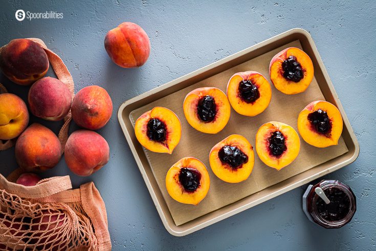 Baking tray with eight peach halves with black cherry jam in the center, in the frame, you will see a bag on the left side with fresh peaches and on the right side an open jar of jam. recipe at spoonabilities.com