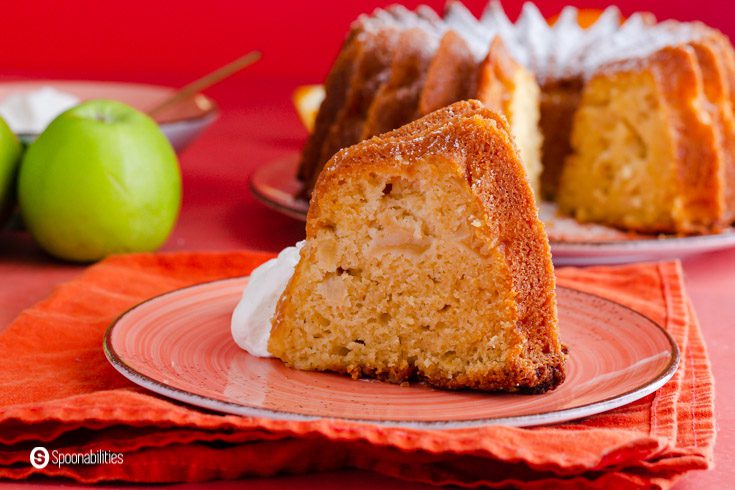 A piece of apple cake made out of olive oil and Granny Smith apples. The slice of cake is on a small plate on top of an orange napkin. Recipe at Spoonabilities.com