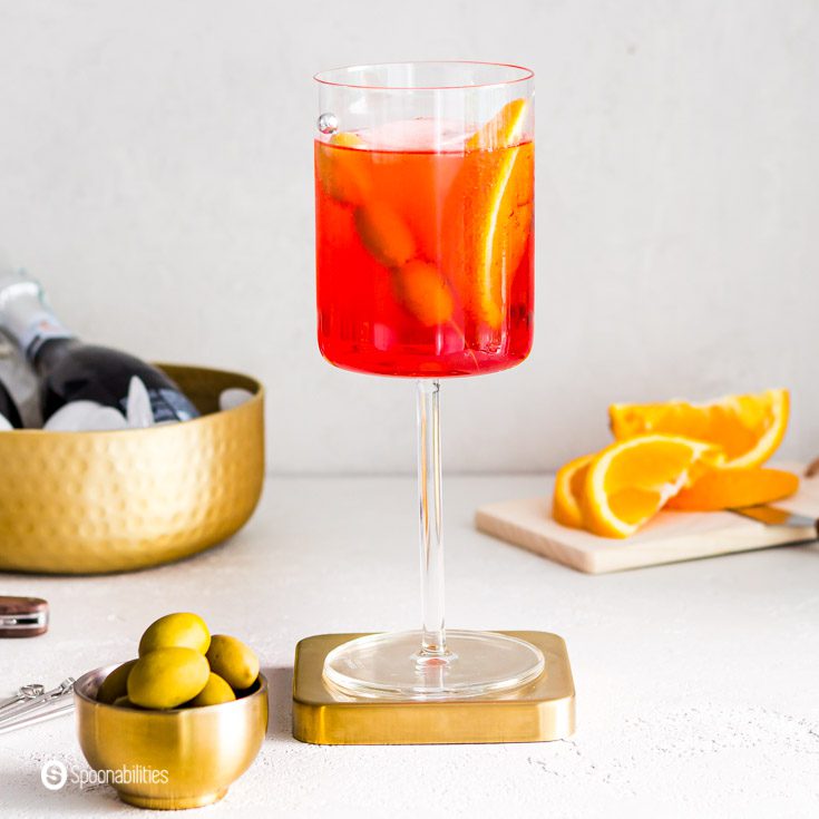 A close up of the famous Italian cocktail Aperol Spritz served in a glass wine. The glass is on top of a golden metal coaster. In a small bowl some green Greek olives. recipe at Spoonabilities.com