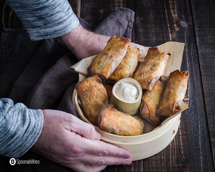Two hands holding a basket with air fryer egg rolls and yogurt sauce. These egg rolls are vegan and baked in the air fryer. Recipe at Spoonabilities.com