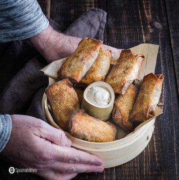 Two hands holding a steam basket with some vegan egg rolls cooked in the air fryer. In the center is a little container with garam masala yogurt sauce. Recipe at Spoonabilities.com