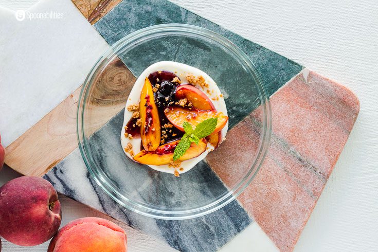 A photo with white background with a marble serving board, on top of the serving board, a glass plate with a restaurant-style presentation with baked peaches, crème Fraiche, black cherry preserves, and garnished with crushed amoretti cookies and fresh mint. Recipe at Spoonabilities.com