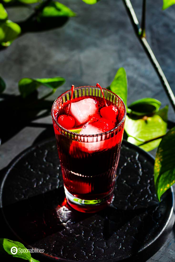 Overhead close-up of a tall glass with a margarita with cherry flavor. The glass is on top of a black platform plate and surrounds plant leaves. Find out more about these tart cherry margaritas at Spoonabilities.com