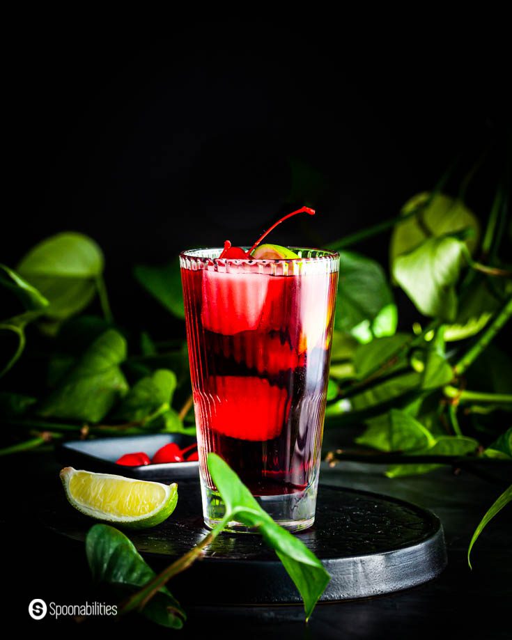 A tall glass on top of a platform. The glass has a margarita cocktail with cherry tart juice, triple sec, and silver tequila and garnished with a maraschino cherry and a lime wedge. Recipe at Spoonabilities.com