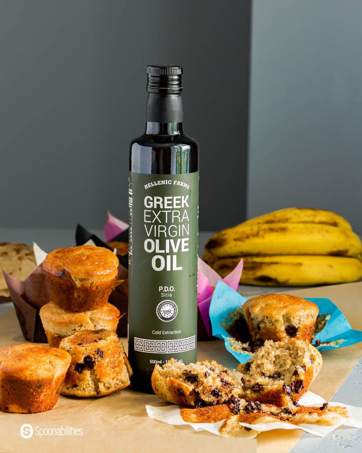 A bottle of Hellenic Farms Extra Virgin Olive oil. The bottle placed in the center of the photo and around the olive oil are sourdough muffins made with bananas and chocolate chips. Recipe at Spoonabilities.com