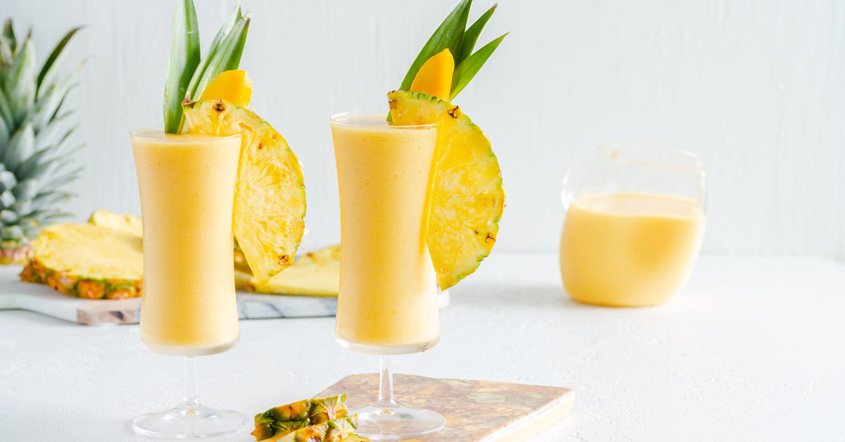 Two tall concave glasses filled with mango pina colada garnished with pineapple leaves and slices