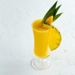 a tall glass of mango pina colada garnished with pineapple leaves and slice