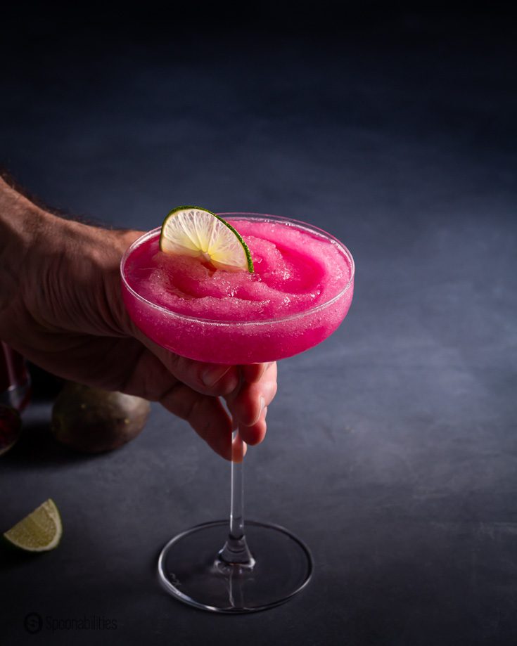 A hand holding a coupe glass with a frozen margarita flavored with prickly pear syrup. Recipe at Spoonabilities.com