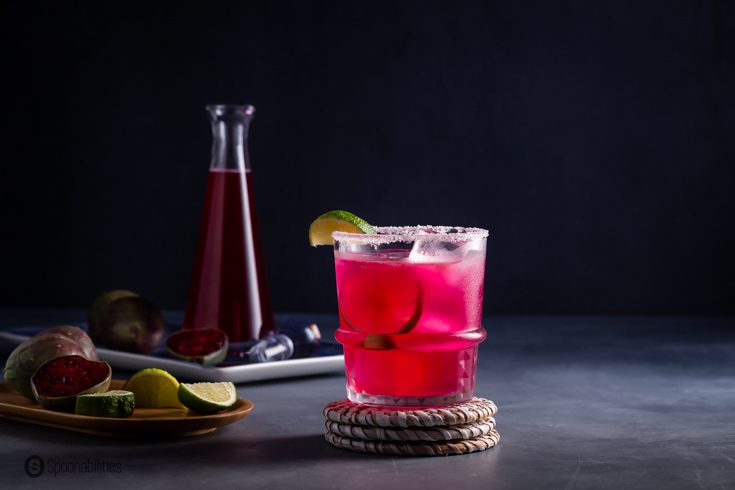 A rock glass on a wicker coaster with a prickly pear margarita. In the background a bottle with the prickly pear syrup and prickly pear cactus fruit. Recipe at Spoonabilities.com