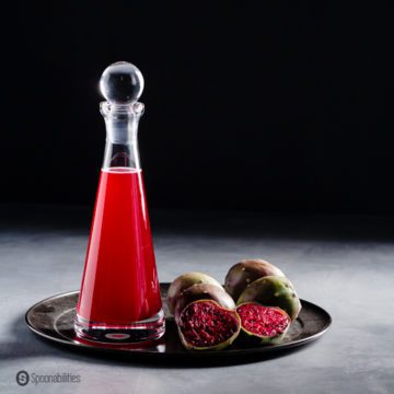 A syrup bottle with prickly pear syrup on a metal tray and next to the bottle are prickly pears fruit. Recipe at Spoonabilities.com