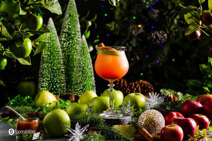 A glass with a Spiked Caramel Apple Cider in the center of the photo. The background is moody and decorated with apples, and Christmas décor.