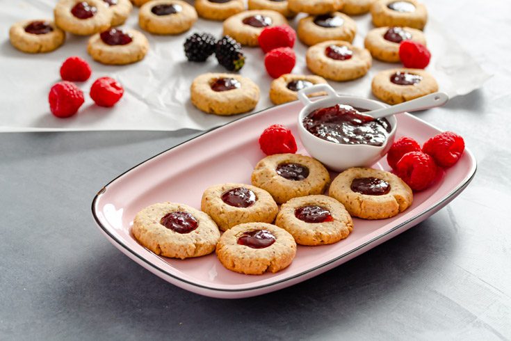 A pink rectangular plate with some almond thumbprint cookies filled with berry jam. On the plate there is a small dipping bowl with some raspberry jam.
