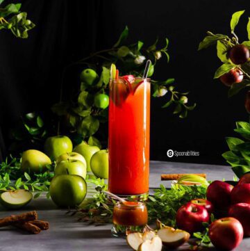 A glass filled with ice, Caramel Apple Cider Vodka Punch, apple slices, and a stick of cinnamon. Around the glass red and green apples, and a dark background.