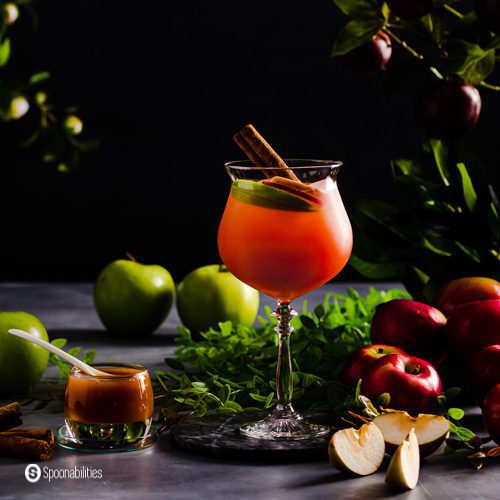 A Tall glass filled with ice, Caramel Apple Cider Vodka Punch, apple slices, and a stick of cinnamon. Around the glass red and green apples, a small cup with caramel sauce, and a dark background.