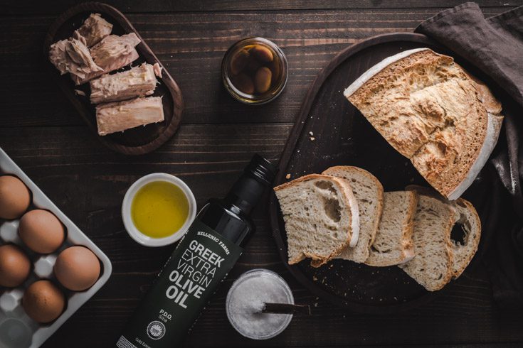 A Bottle of Hellenic Farms Olive oil and pieces of bread, eggs, salt, and tuna around the bottle of oil.