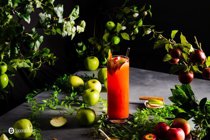 A large pitcher with a punch made with vodka, caramel sauce, grenadine, apple cider, and lemon juice. Also, slices of green and red apple. Surrounding the pitcher branches of apple tree and some apples on the floor.