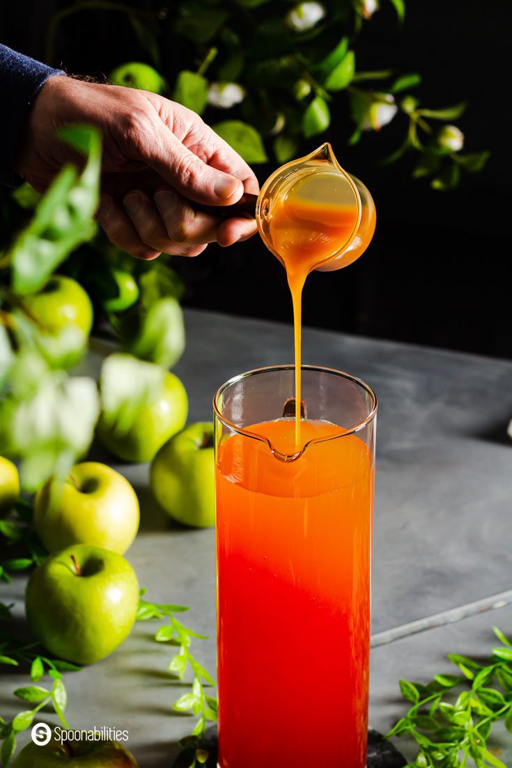 A hand pouring caramel sauce into a pitcher with apple cider punch. In the background you can see some green apples on the table.
