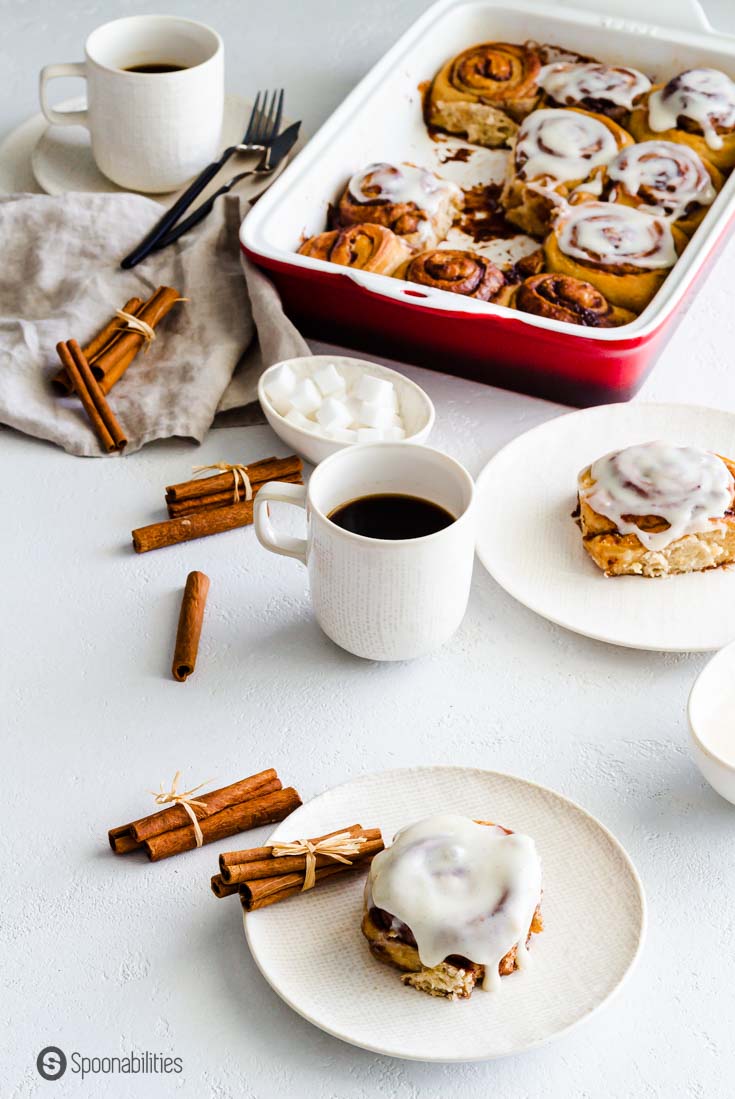 A table with cinnamon rolls made with sourdough in a red baking dish. A couple of small plates with one serving of the rolls.