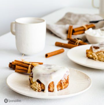 White plate with one Sourdough Cinnamon Rolls. In the background more cinnamon buns and cups with coffee.