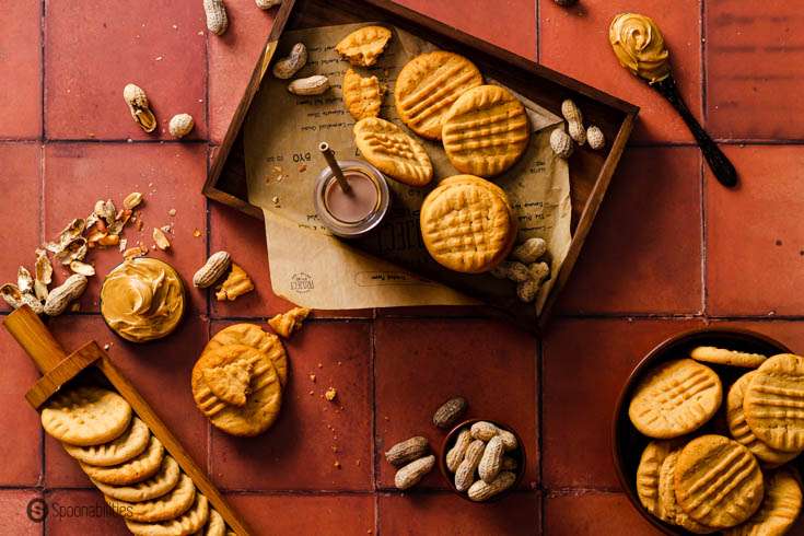 Terracotta tiles tabletop with some peanut butter cookie, peanut in shell, peanut butter in a bowl, and a hot chocolate in a bottler.