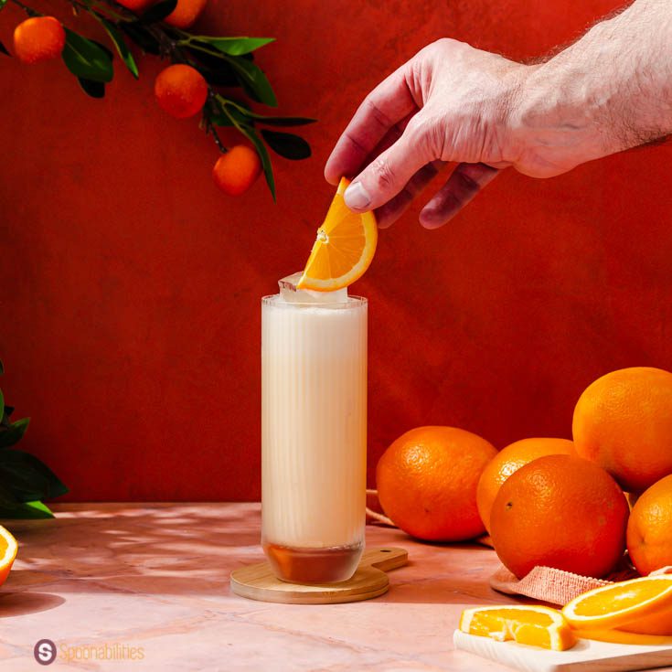 A hand garnishing a highball glass with an orange slice to the Dominican drink Morir soñando with alcohol.