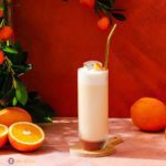 A tall glass in the center of the photo with drink Orange Creamsicle Cocktail. In the background some oranges.