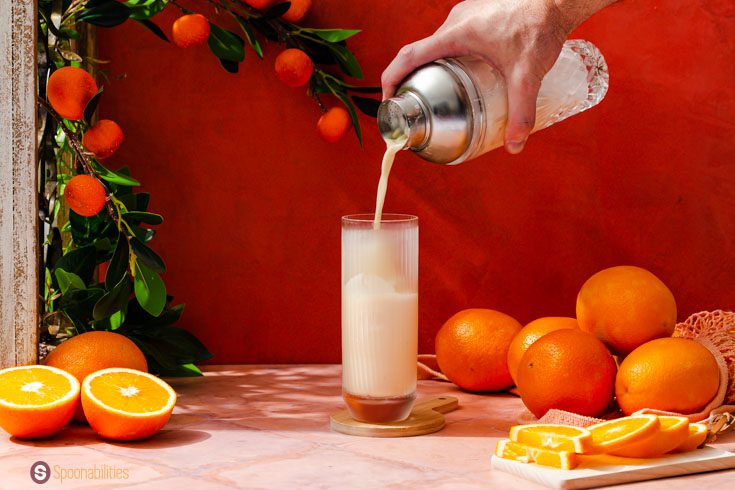 A hand pouring the the Creamsicle Drink with Gin in a tall glass. In the background some oranges.