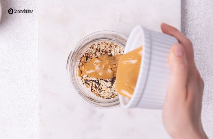 Hand pouring almond butter from a white ramekin into a glass jar of oats mixture.