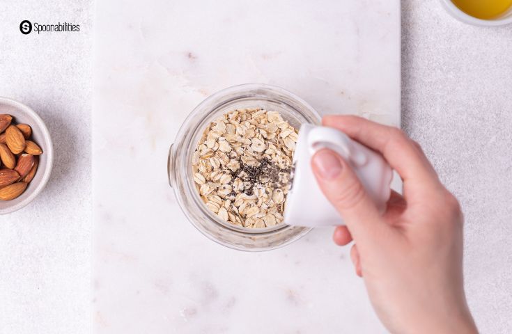 Hand pouring chia seeds into a glass container of old-fashioned rolled oats
