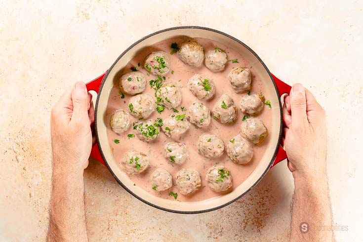 Dutch oven pot of Swedish Meatballs swimming in lingonberry gravy being held by two hands