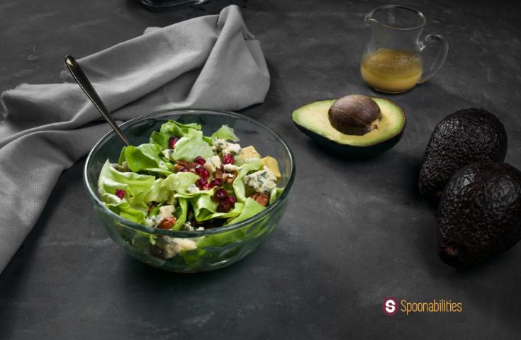 a photo of Honey mustard vinaigrette on butter lettuce pear salad in a clear bowl with a fork, a glass of honey, avocados, and gray cloth on the side