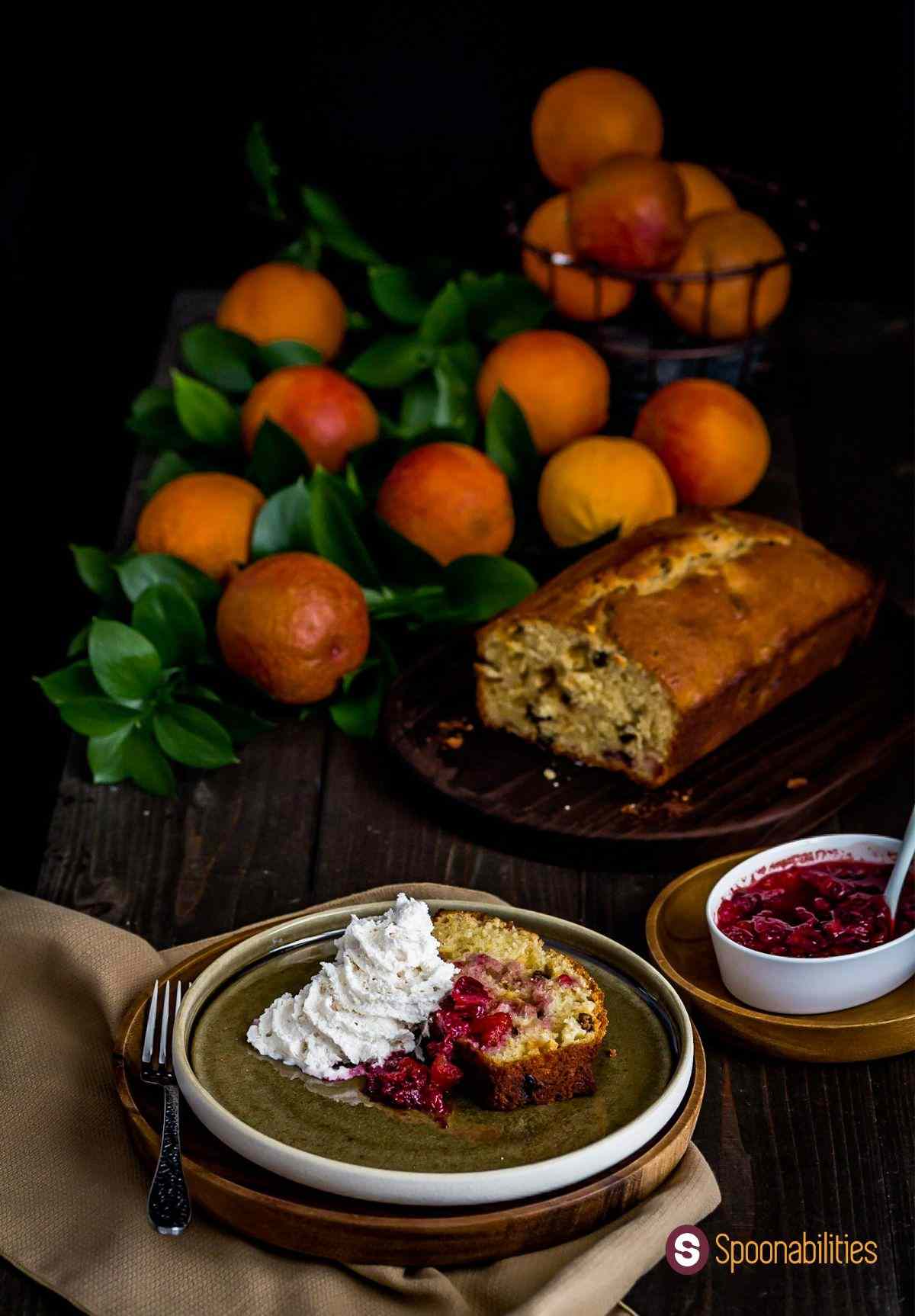 A slice of Blood Orange olive oil cake in the front of the photo on a plate, with compote on the side. Several blood oranges scattered in the background. More gourmet recipes at Spoonabilities.com