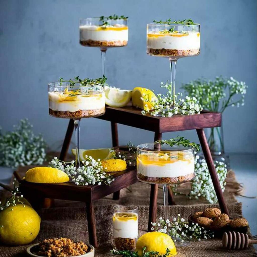 Display of no bake honey cheesecakes in dessert glasses with lemons, white flowers and amaretto cookies