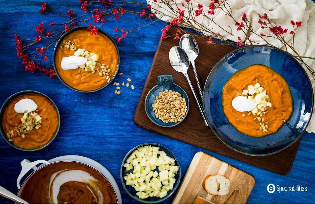 Pumpkin bisque in three blue bowls with diced apples and pumpkin seeds and spoons