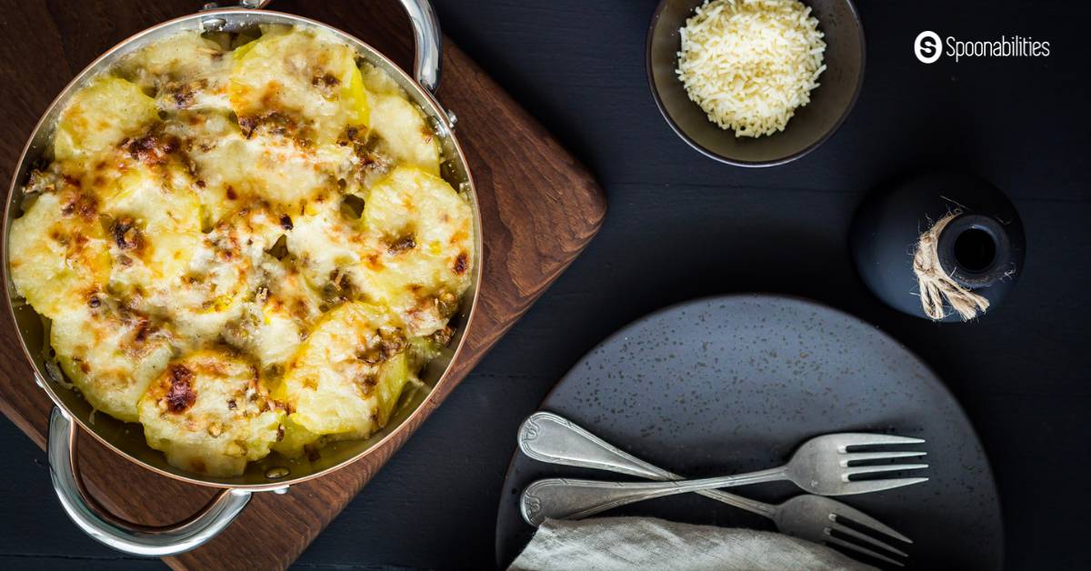 Leek and potato gratin in a stainless pot with cheese topping in a small bowl and a black plate with two forks at the side